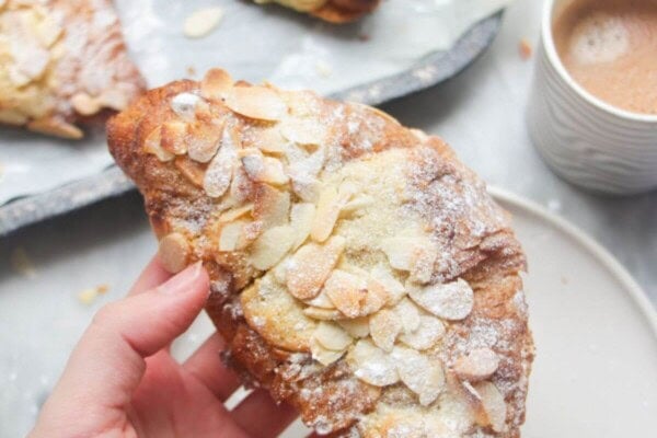 A hand holding an almond croissant with more almond croissants in the background.