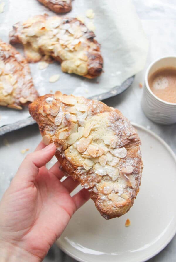 A hand holding an almond croissant with more almond croissants in the background.