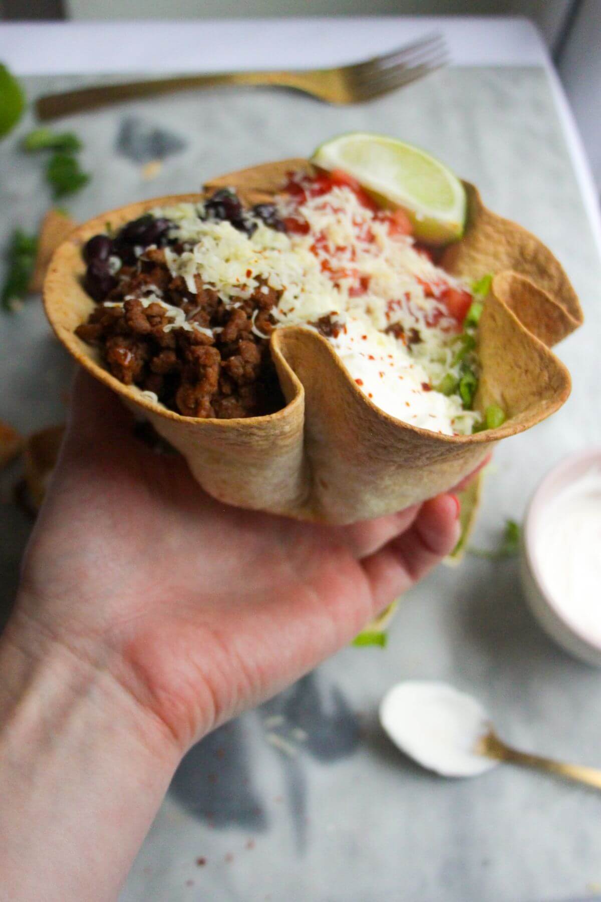 Hand holding loaded taco salad bowl with small jar of yogurt in the background.