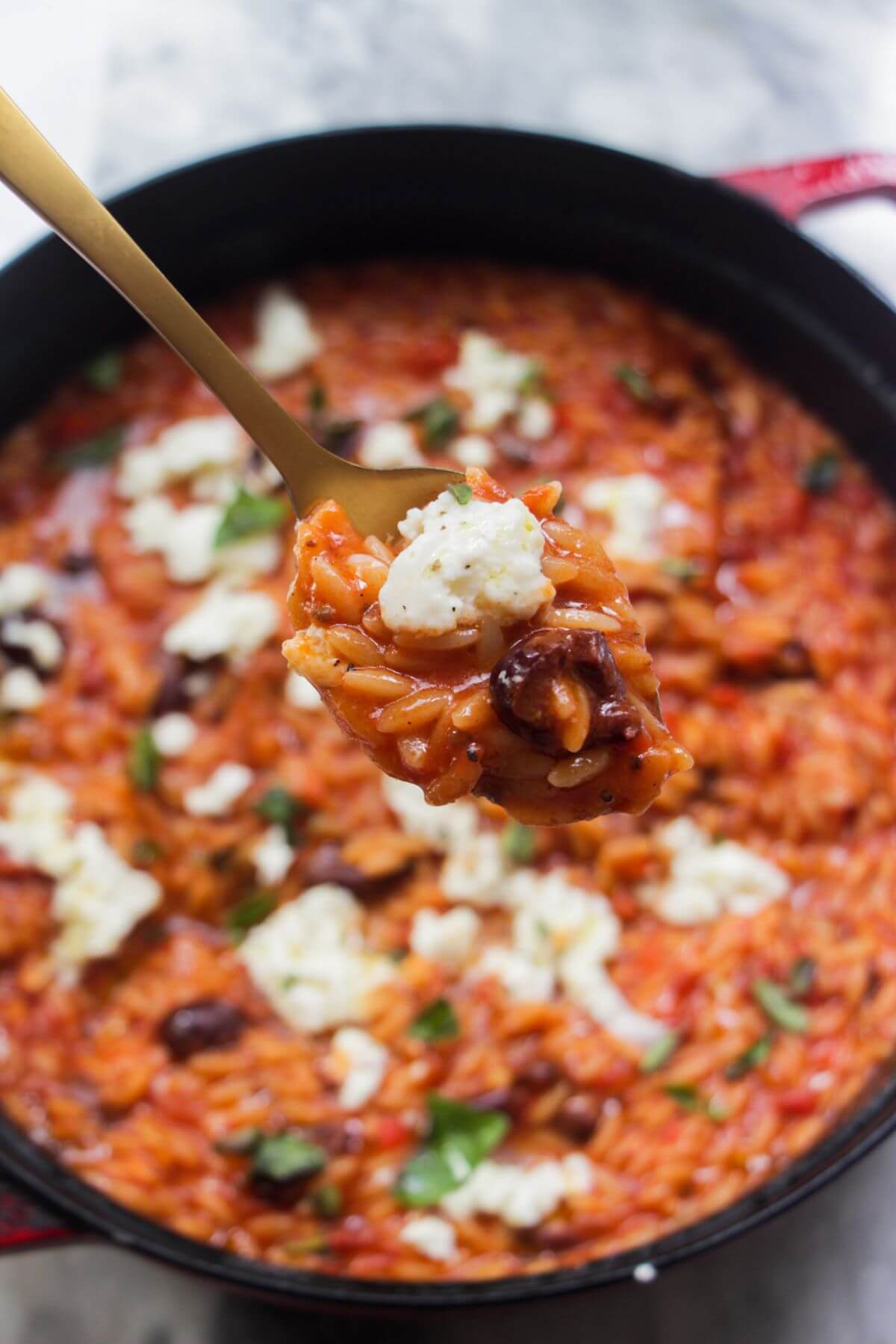 Close up of tomato orzotto on a small spoon with the rest of the orzo in the backgorund in a large skillet.