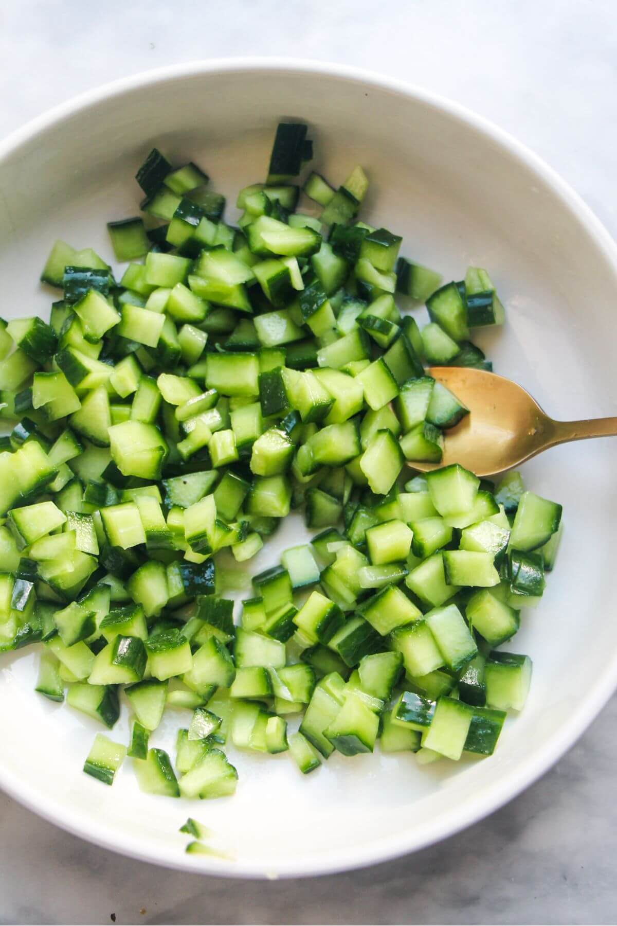 Small white bowl filled with diced cucumber with a gold spoon mixing it.