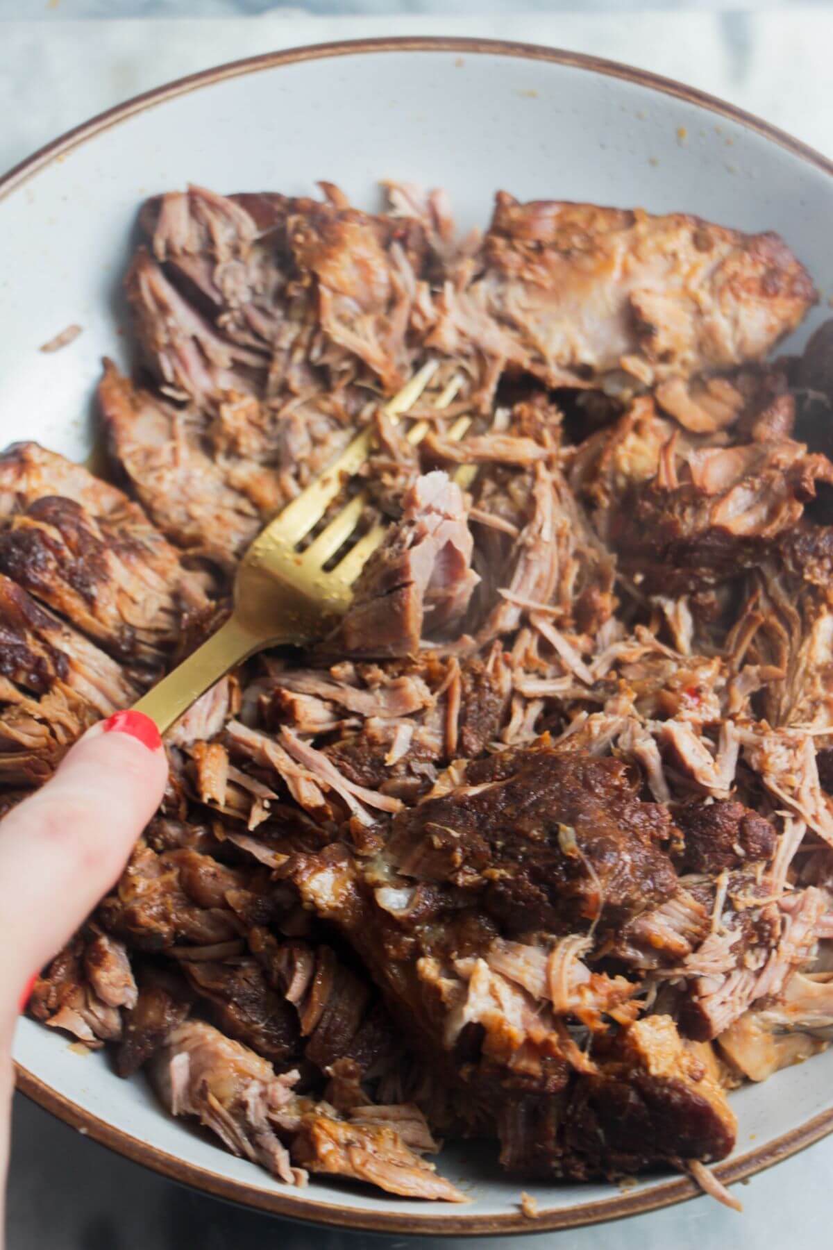 Hand holding a gold fork pulling pork in a bowl.