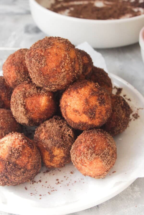 A pile of sugar coated donut bites on a small white plate, with bowl of more pumpkin spice sugar in the background.
