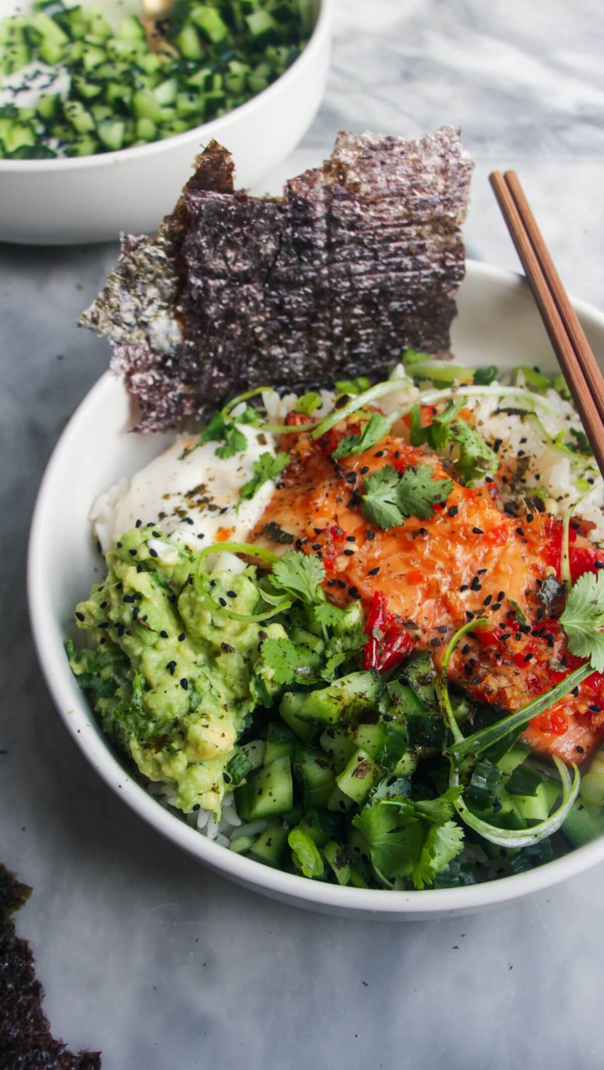 White bowl filled with colourful vegetables and a salmon fillet with seaweed sheets on the side and chopsticks in the bowl.