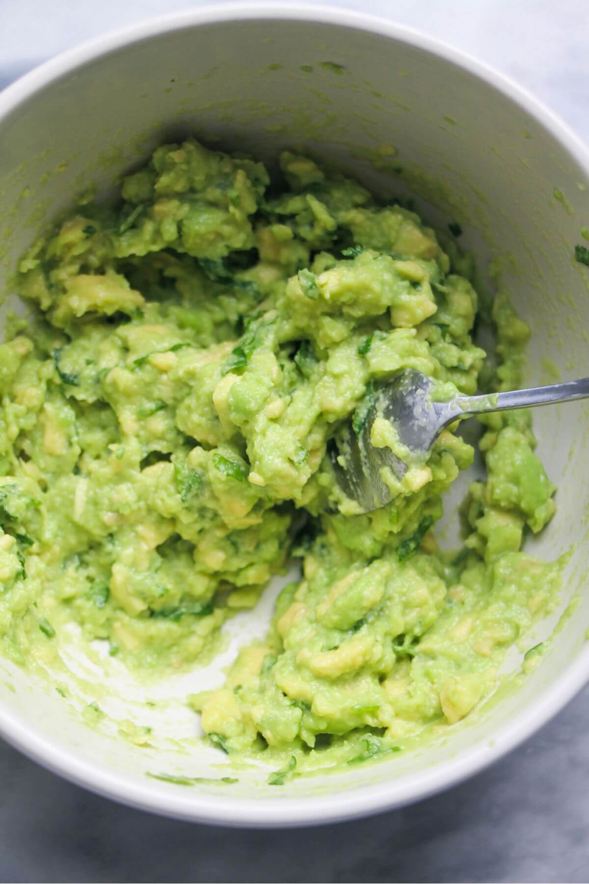 Small silver spoon mixing green avocado into a smooth mash in a small white bowl.