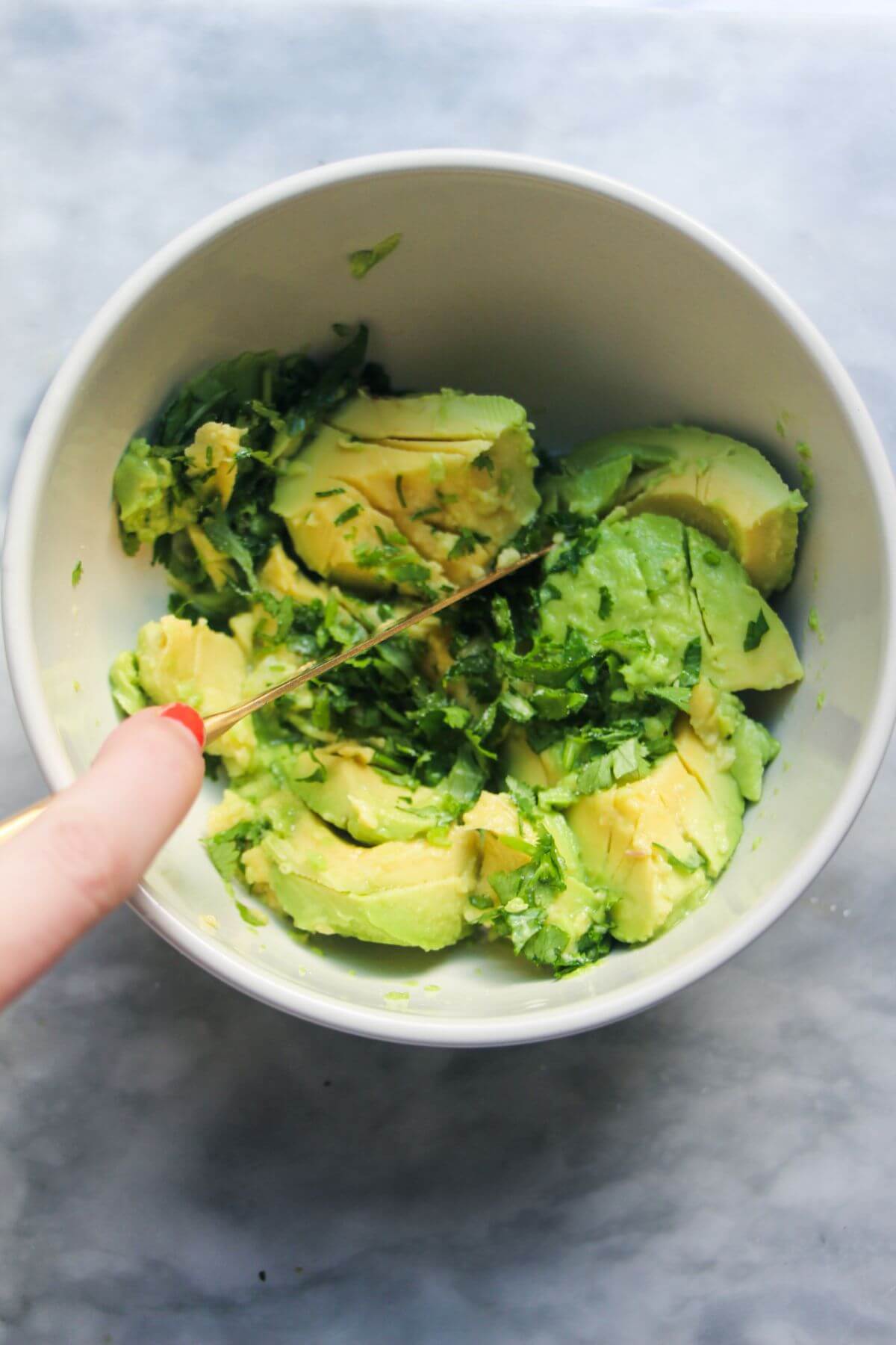 Avocado flesh and chopped coriander in a small white bowl with a hand holding a gold knife mixing them.