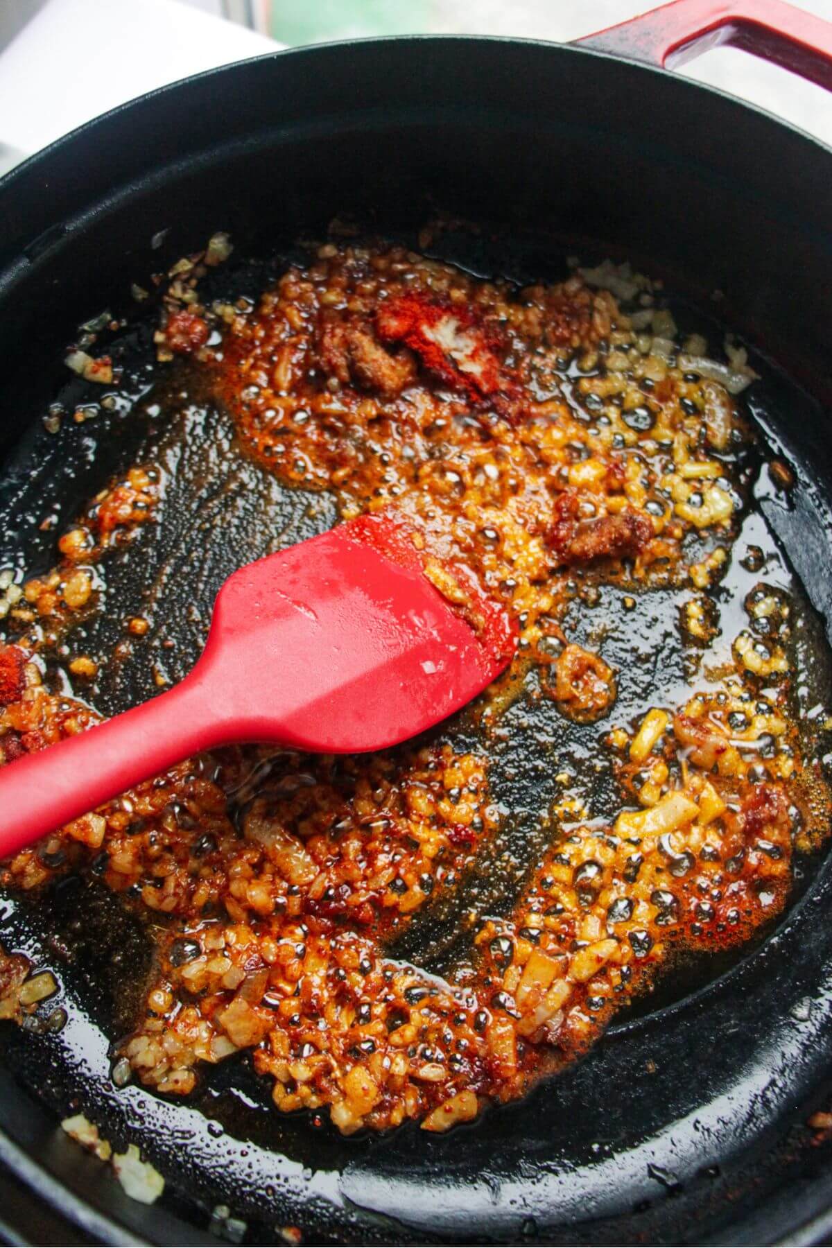 Red spatula stirring spices through diced onion and garlic in a black pan.