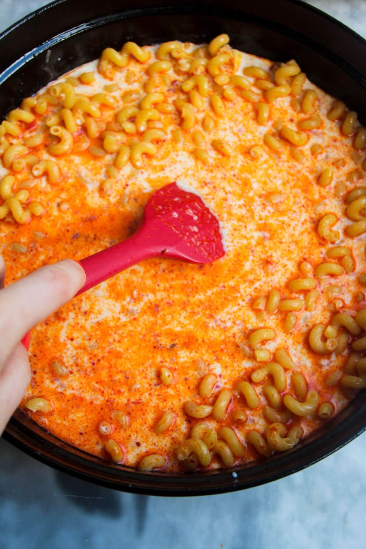 Red spatula stirring mac and cheese in a large black skillet.