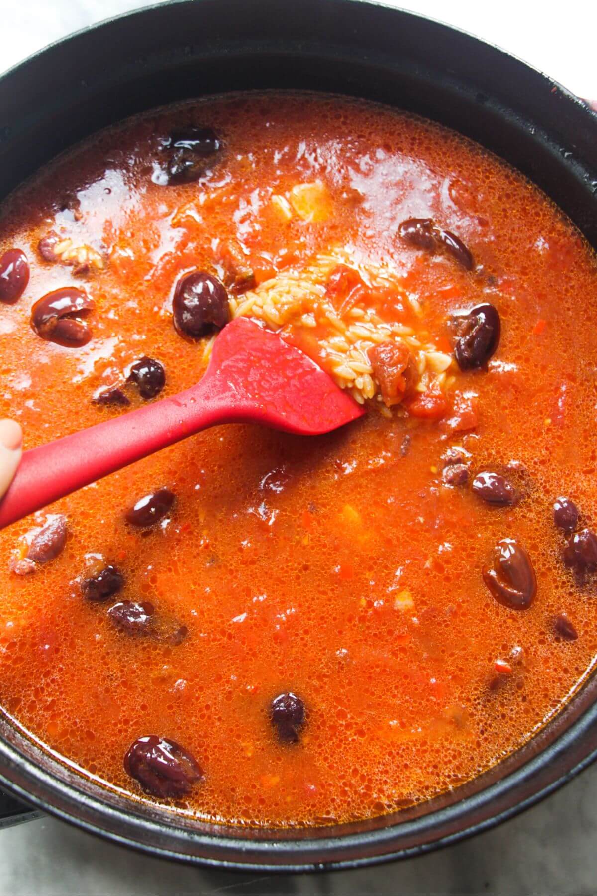 Red spatula stirring stock through tomatoes, orzo and olives in a large black skillet.