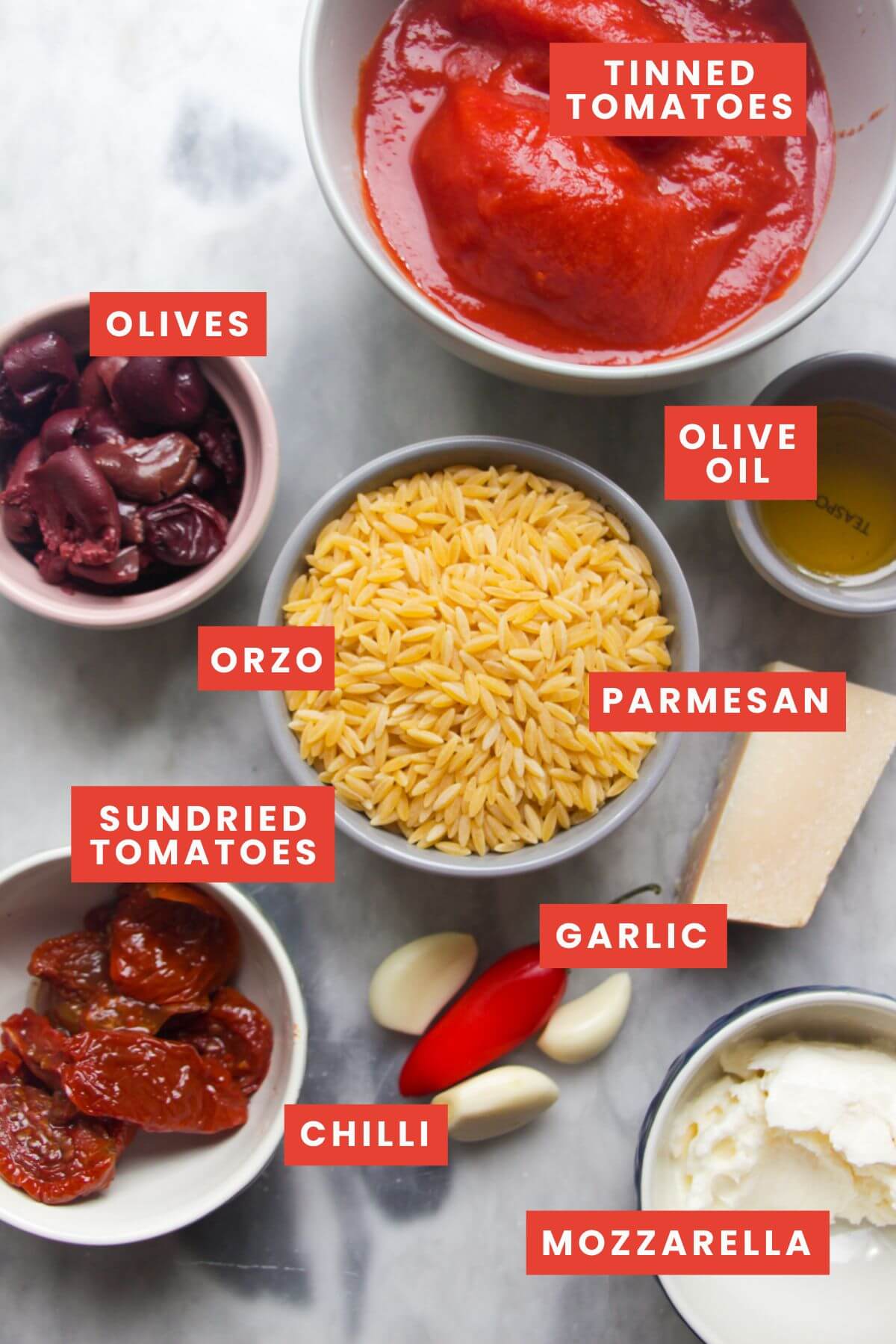 Ingredients for cheesy tomato orzotto laid out on a grey marble background and labelled.