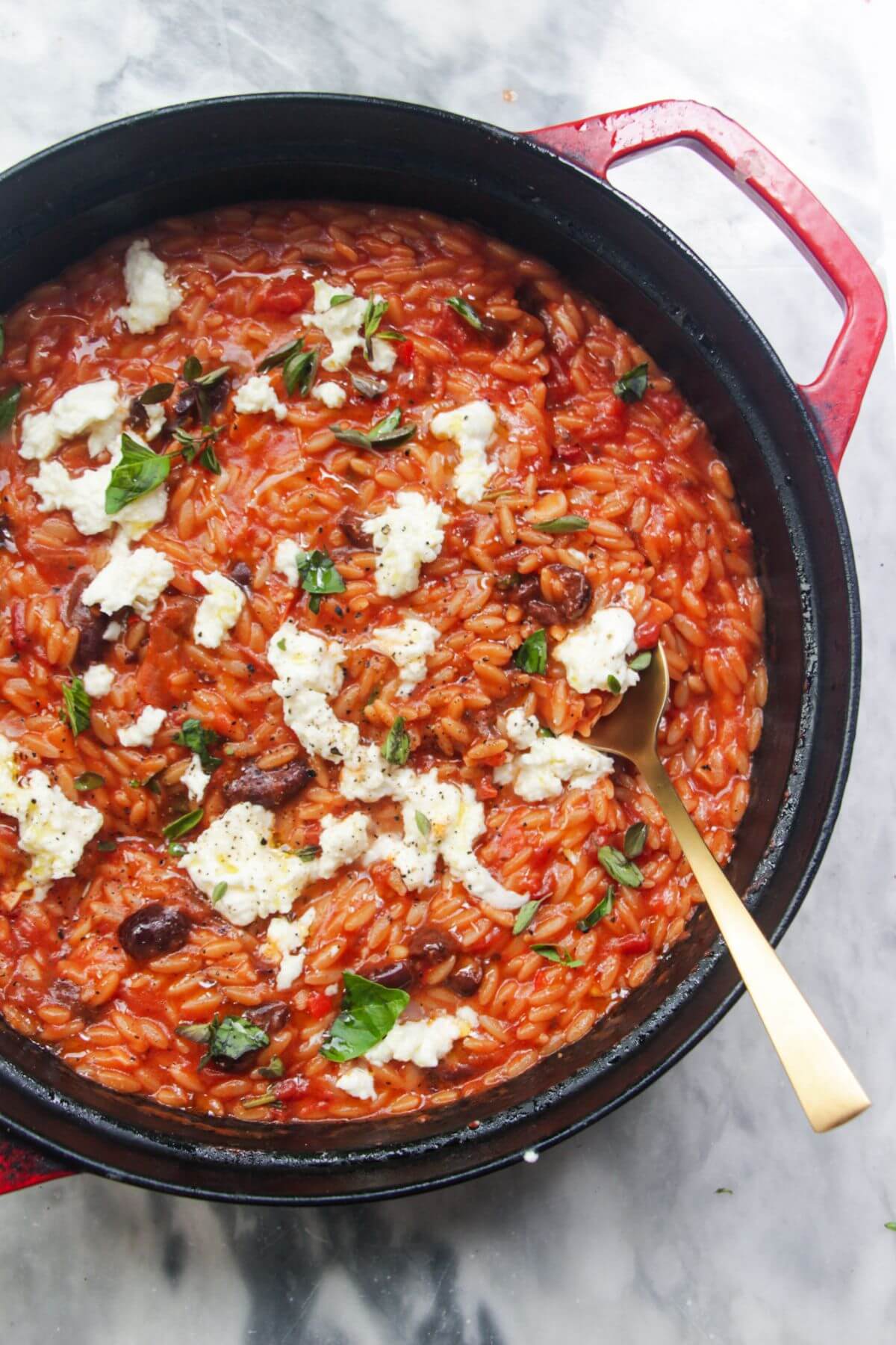 Cheesy tomato orzotto in a large black and red skillet on a grey marble background.