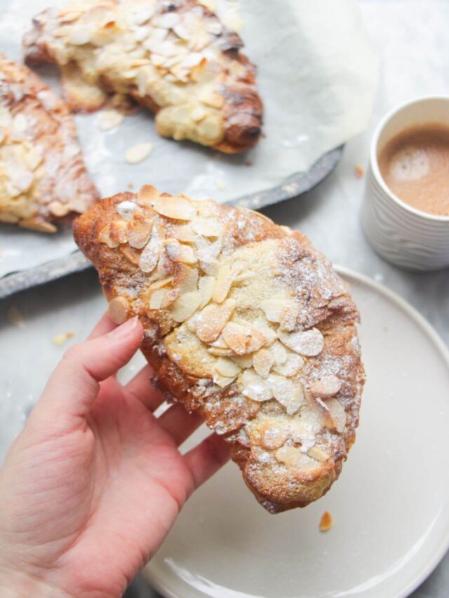 A hand holding an almond croissant with more almond croissants in the background.