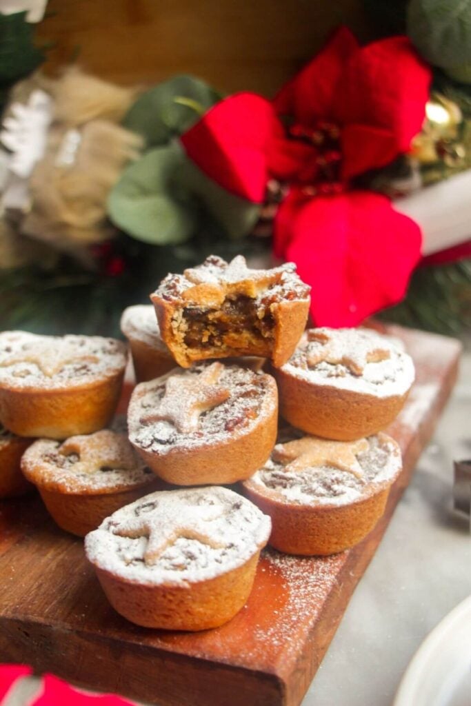 A pile of icing sugar dusted Christmas mince pies on a small wooden board with Christmas deocrations in the background.