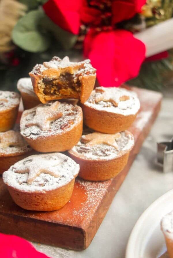 A pile of icing sugar dusted Christmas mince pies on a small wooden board with Christmas deocrations in the background.