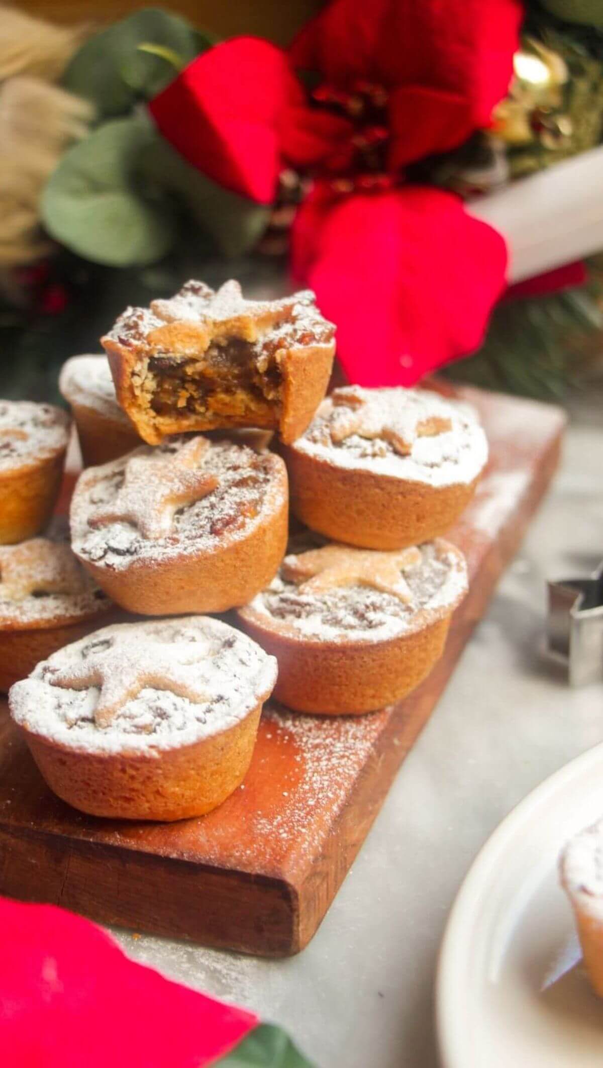 A pile of icing sugar dusted Christmas mince pies on a small wooden board with Christmas deocrations in the background.