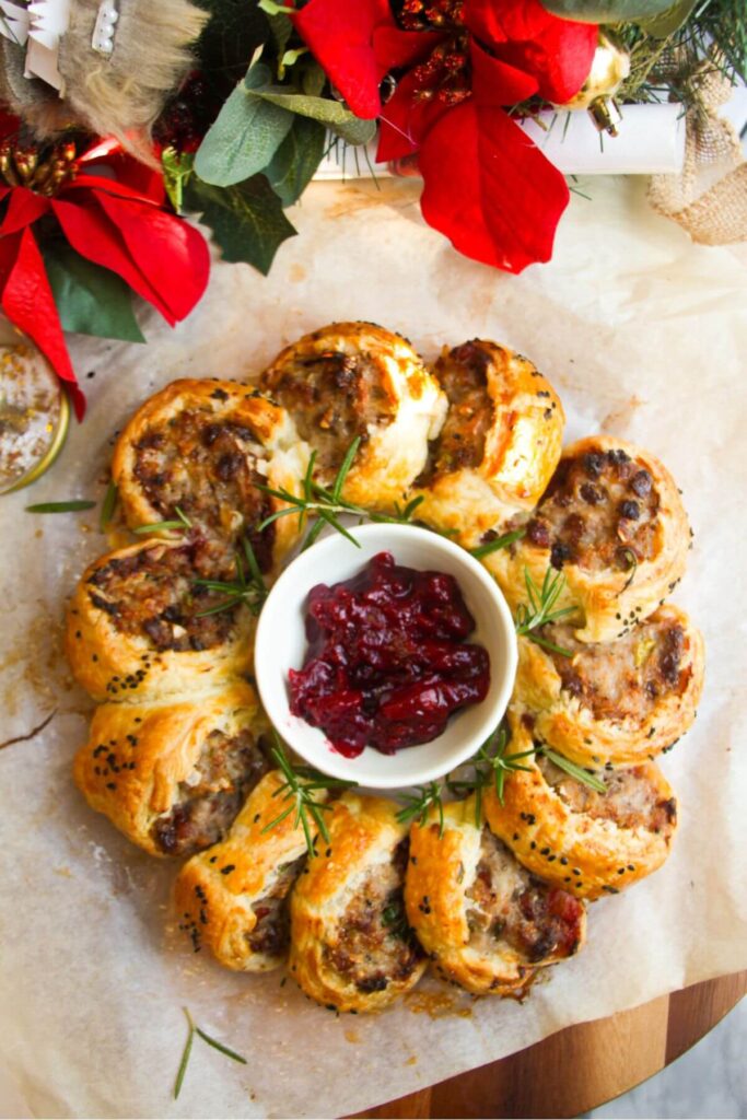 Christmas sausage roll wreath on baking paper on a wooden board with Christmas decorations in the backgorund.