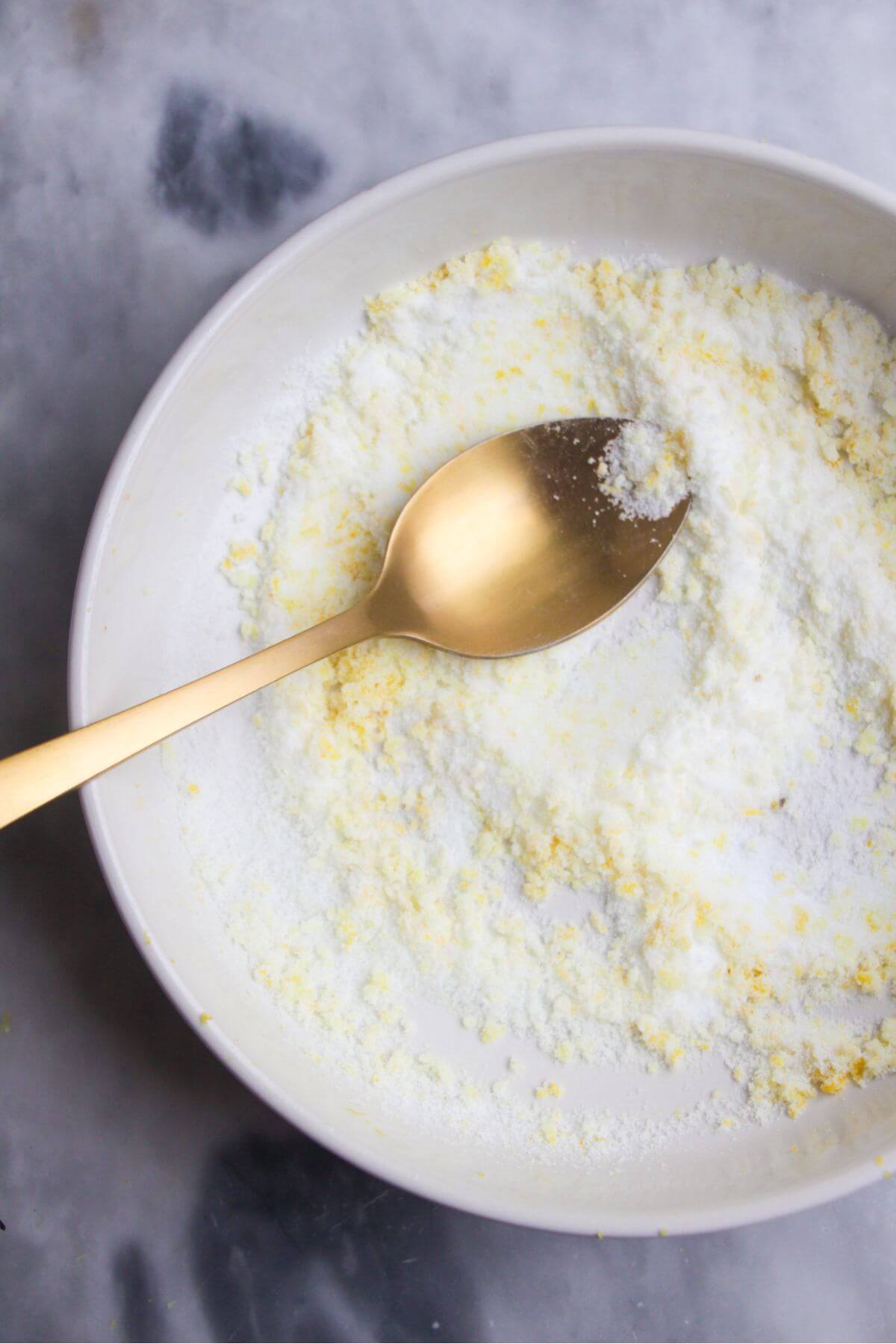 Gold spoon mixing salt and lemon zest in a smal white bowl.