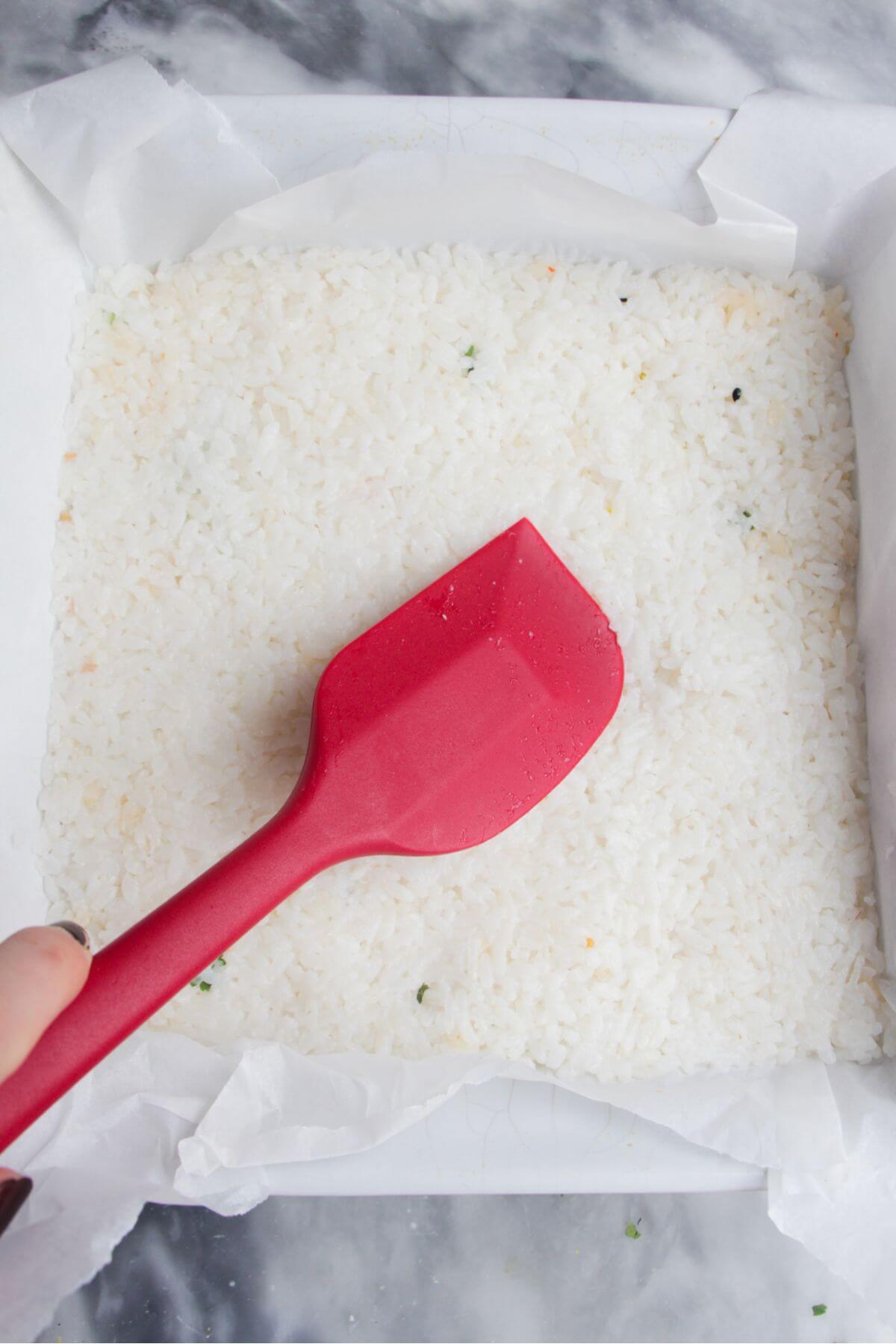 Red spatula smoothing rice into a square baking dish.