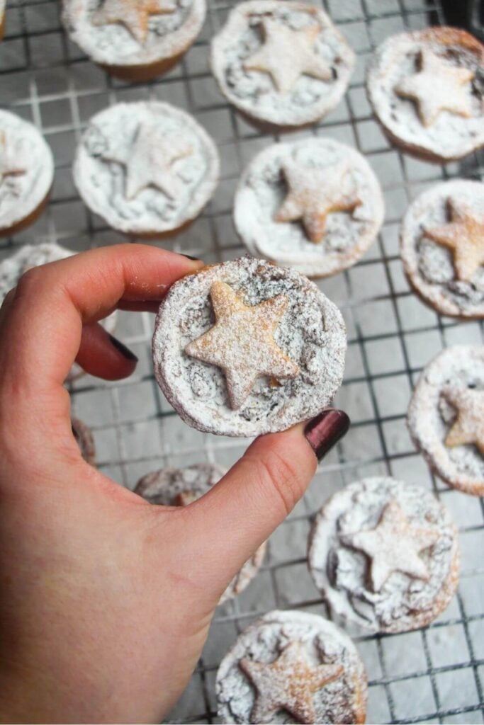 A hand holding a Christmas fruit mince pie dusted with icing sugar with more mince pies in the background.
