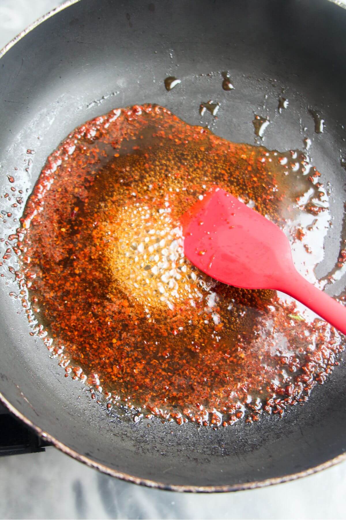 Honey and chilli flakes bubbling up in a small black pan, being stirred with a red spatula.