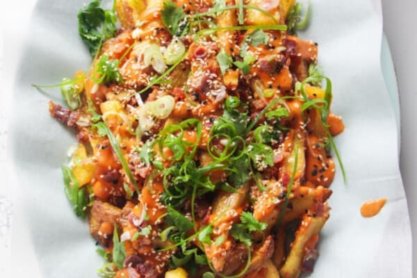 Loaded potato wedges on a large oval white plate with small jug of gochujang bechamel on the side.