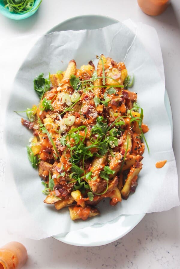 Loaded potato wedges on a large oval white plate with small jug of gochujang bechamel on the side.