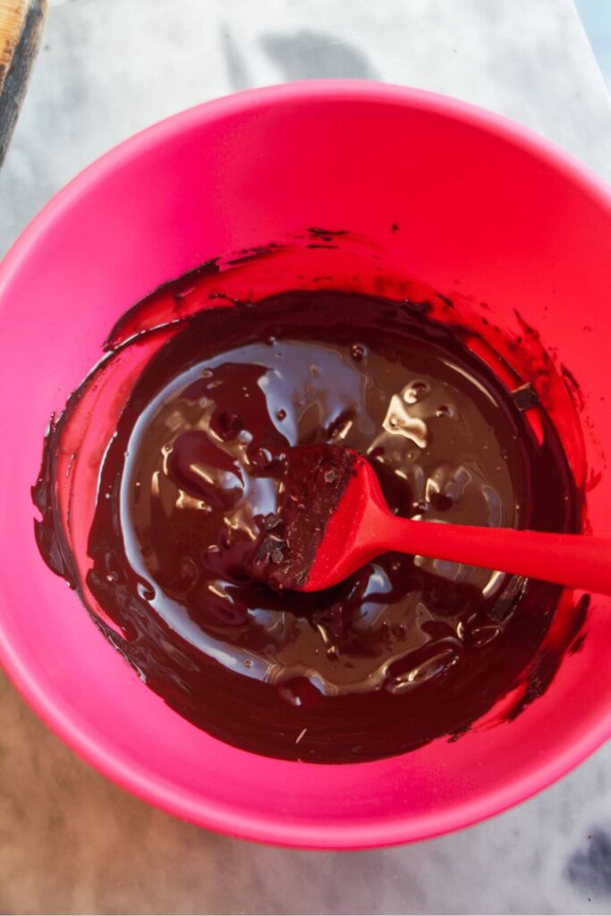 Melted dark chocolate in a pink bowl being stirred with a red spatula.