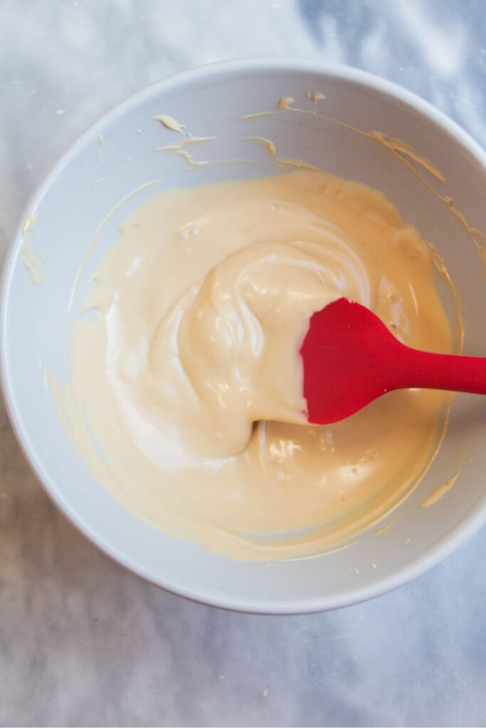 Melted white chocolate in a small white bowl being stirred with a red spatula.