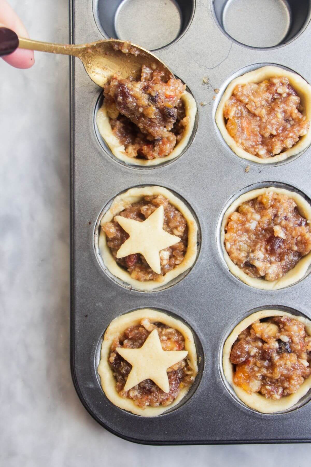 A spoon filling pastry case with fruit mince in a small muffin tin.