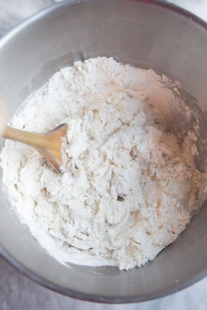 A wooden spatula mixing flour, milk, yeast and salt in a large silver mixing bowl.