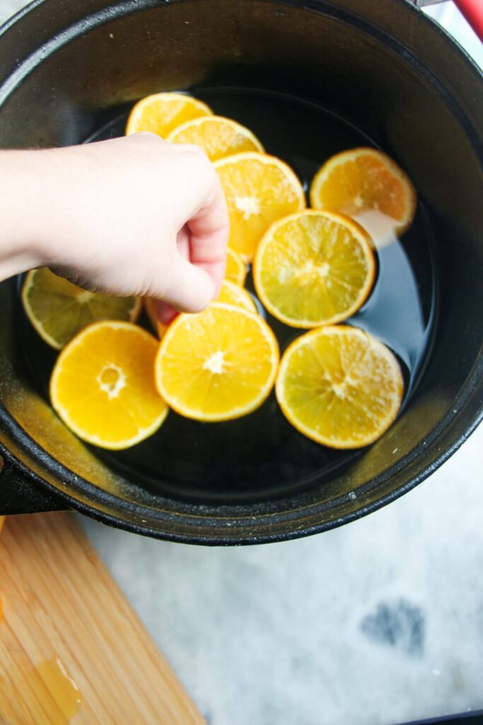 Hand placing orange slices into sugar syrup in a large pot.
