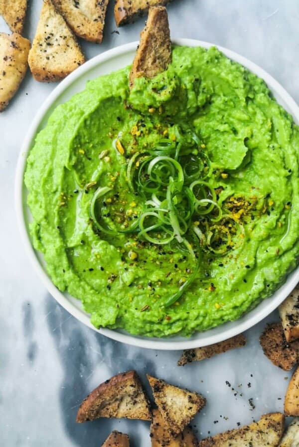Pea and avocado hummus on a small white plate with pita chips on the side.