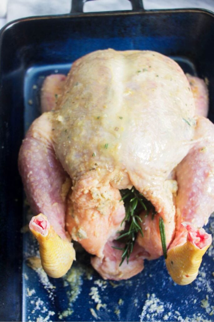 Rosemary sprig in the cavity of a whole chicken in a blue oven dish.