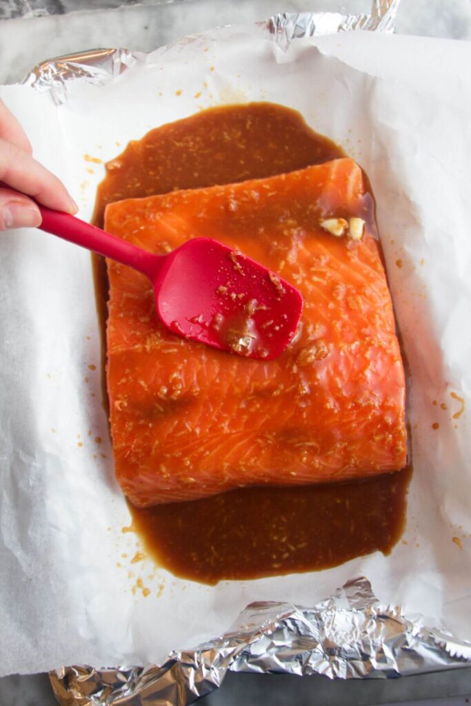 Small red spatula spooning maple miso glaze over a salmon fillet in a baking paper lined tray.