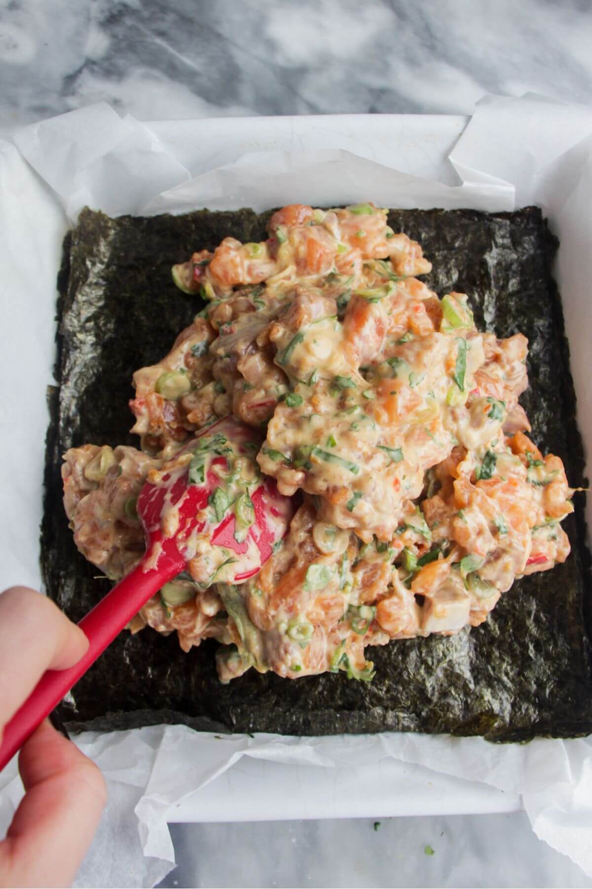 Red spatula spooning salmon sushi mix on top of seaweed sheet in a square baking dish.