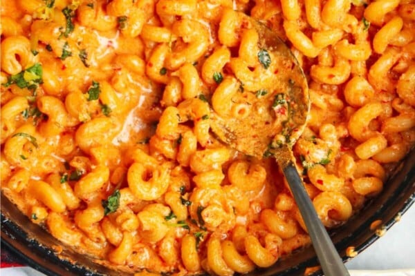 Spicy mac and cheese in a large black skillet with a silver spoon inside and pink plate next to it.