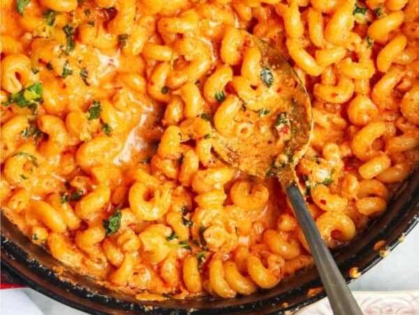 Spicy mac and cheese in a large black skillet with a silver spoon inside and pink plate next to it.