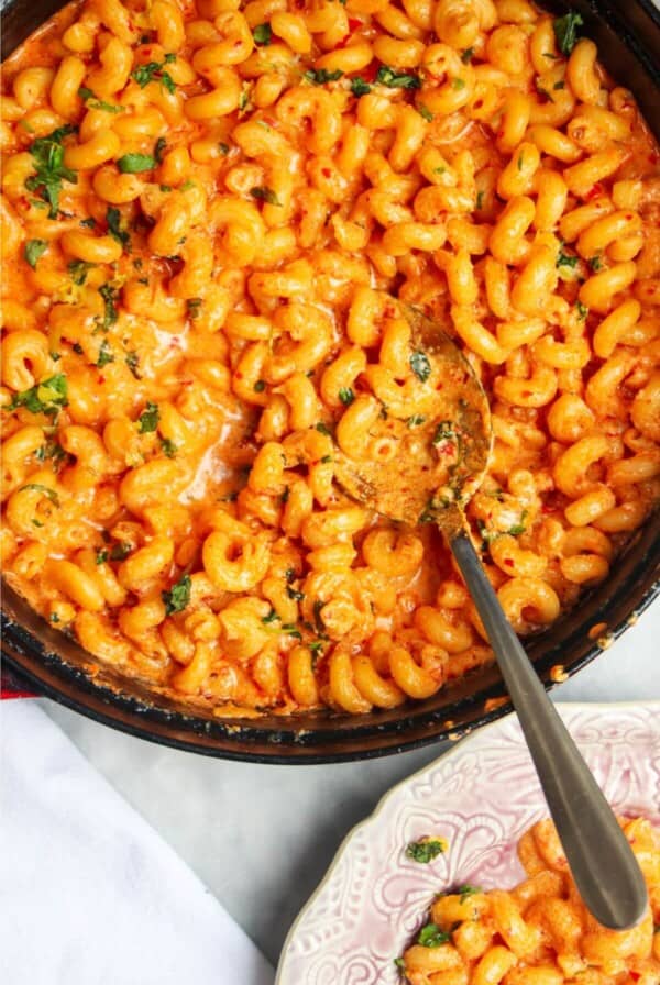 Spicy mac and cheese in a large black skillet with a silver spoon inside and pink plate next to it.
