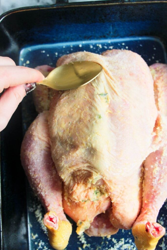 Gold spoon pressing butter down underneath the skin of a chicken in a blue dish.