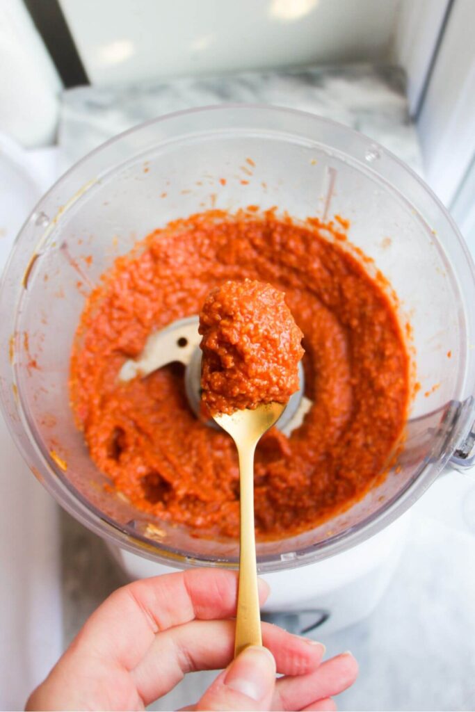 Tomato pesto on a small gold teaspoon with more pesto in a food processor in the background.