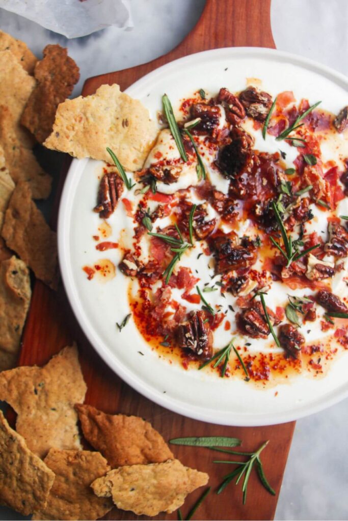 Whipped feta dip topping with hot honey and crispy prosciutto shards on a white plate with crackers on the side.