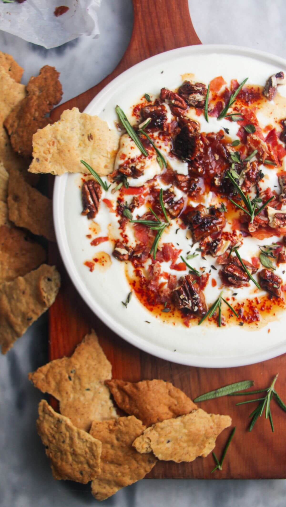 Whipped feta dip topping with hot honey and crispy prosciutto shards on a white plate with crackers on the side.