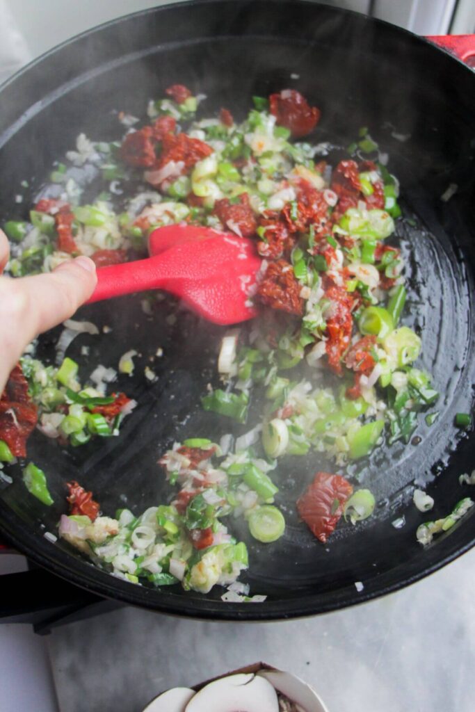 Red spatula stirring chopped sundried tomatoes through spring onions and shallots in a large black skillet.