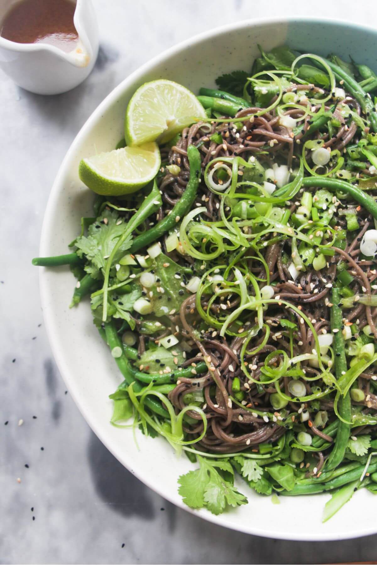 Cold Asian soba noodle salad with lots of green vegetables in a large serving platter, with jug of dressing on the side.