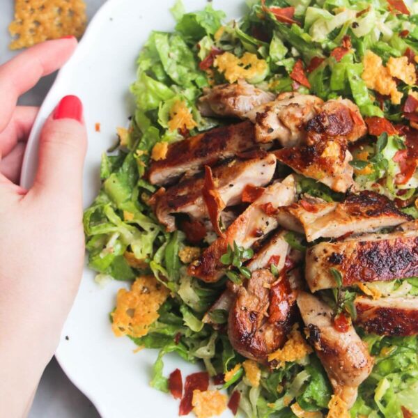 A hand holding a large white plate with sliced grilled chicken on top of green chopped lettuce.