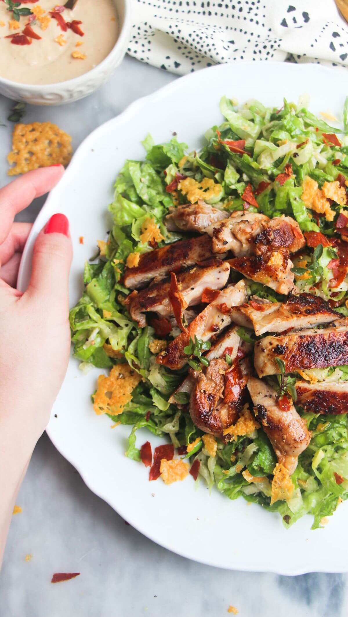 A hand holding a large white plate with sliced grilled chicken on top of green chopped lettuce.