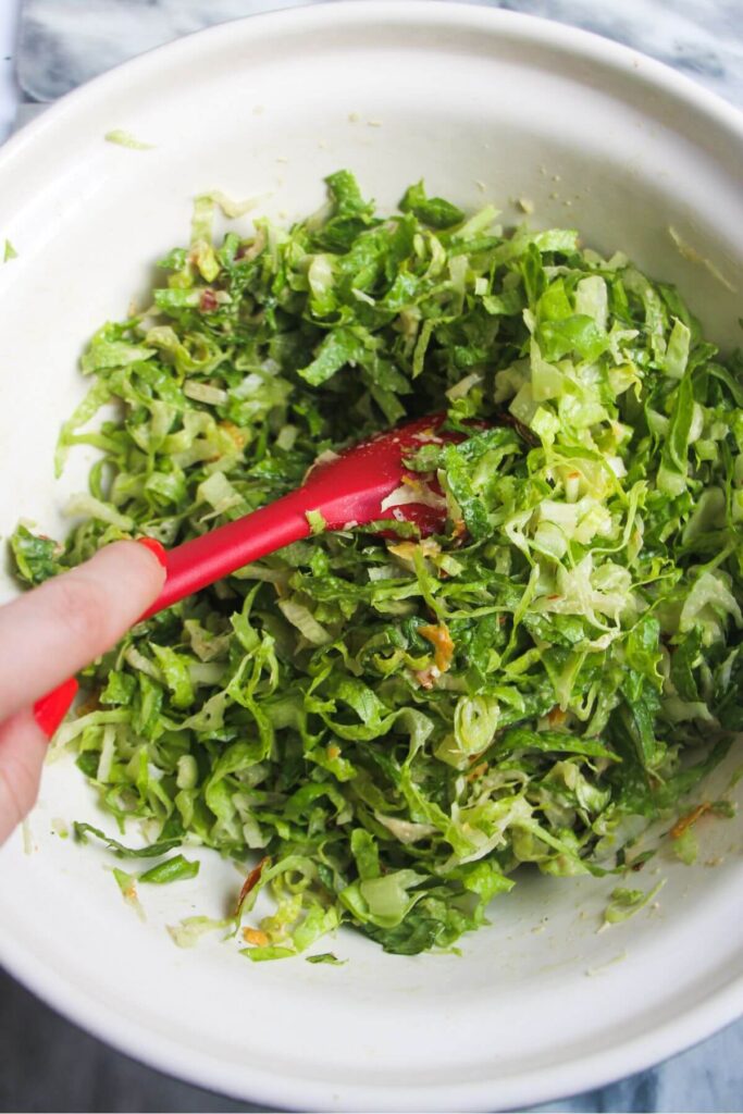 Hand holding a red spatula stirring dressing through lettuce in a large ceramic bowl.