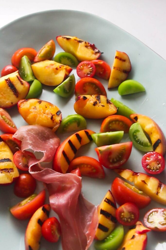 Piece of prosciutto nestled in between sliced tomatoes and grilled peaches on a large oval pale blue dish.