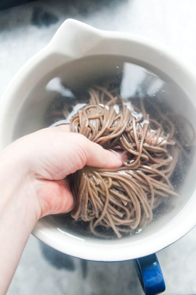 A hand rinsing cooked soba noodles in a bowl of cold water.