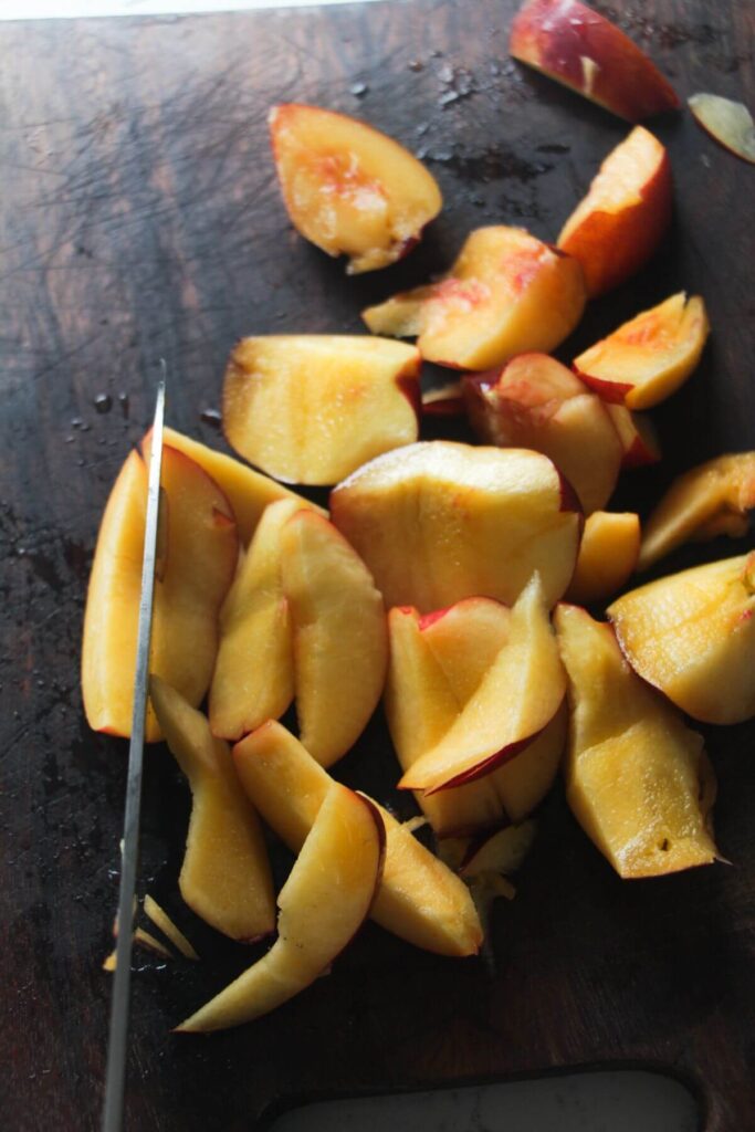 A knife slicing peaches on a dark wooden board.